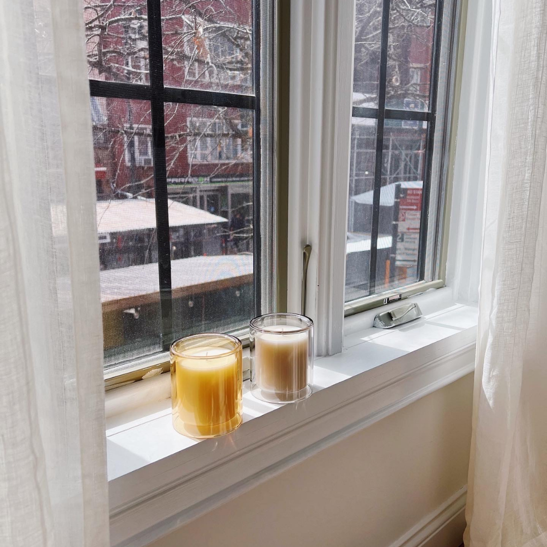 two candles on window stand. one in marigold colored vessel (amber fragrance) and one in dove colored vessel (santal fragance)