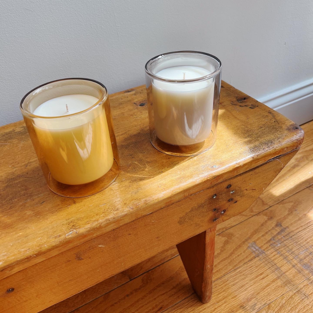 two candles on bench. living room decor. one in marigold colored vessel (amber fragrance) and one in dove colored vessel (santal fragrance)